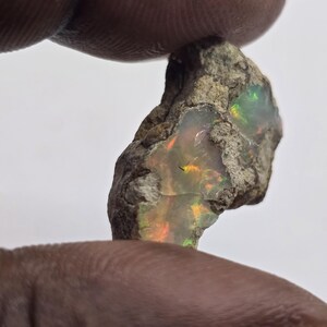 May include: A rough, natural opal gemstone with a matrix of tan and gray rock. The opal displays vibrant flashes of color, including orange, green, and yellow. The gemstone is held between two fingers against a white background.