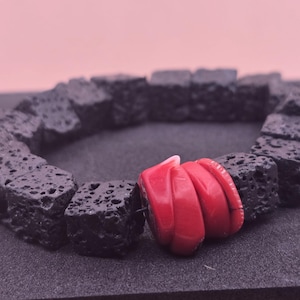 May include: A black lava rock bracelet with a vibrant red coral accent. The bracelet features square-shaped, porous black beads. The coral accent is a cluster of three rounded, red pieces. The bracelet is displayed on a dark surface.