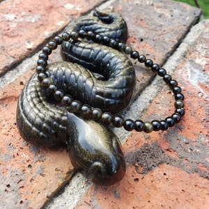 May include: A black and gold beaded necklace made of goldstone beads. The necklace is wrapped around a black and gold snake figurine.