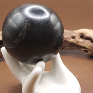 Könnte beinhalten: Eine polierte schwarze Kugel, möglicherweise ein Edelstein, ruht in einem weißen, handförmigen Keramikhalter. Die Kugel hat eine reflektierende Oberfläche. Der Halter ist glatt und glänzend. Der Hintergrund ist warmbraun.