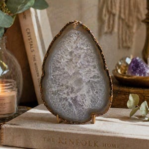 May include: A large, polished agate slice with a gold-colored edge, displayed on a small gold stand. The agate features a white and lavender interior with a crystalline pattern. The slice is displayed on a book with the text "The Kinfolk Home".