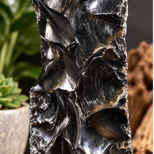 Puede incluir: Una pieza alta y vertical de obsidiana negra con una superficie brillante y reflectante. La piedra presenta fracturas concoideas afiladas y bordes dentados creados por el tallado, lo que resulta en una apariencia texturizada.