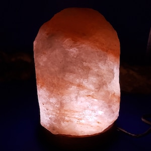 K&ouml;nnte beinhalten: Eine rosa Himalaya-Salzlampe mit einem Holzsockel. Die Lampe ist eingeschaltet und leuchtet.