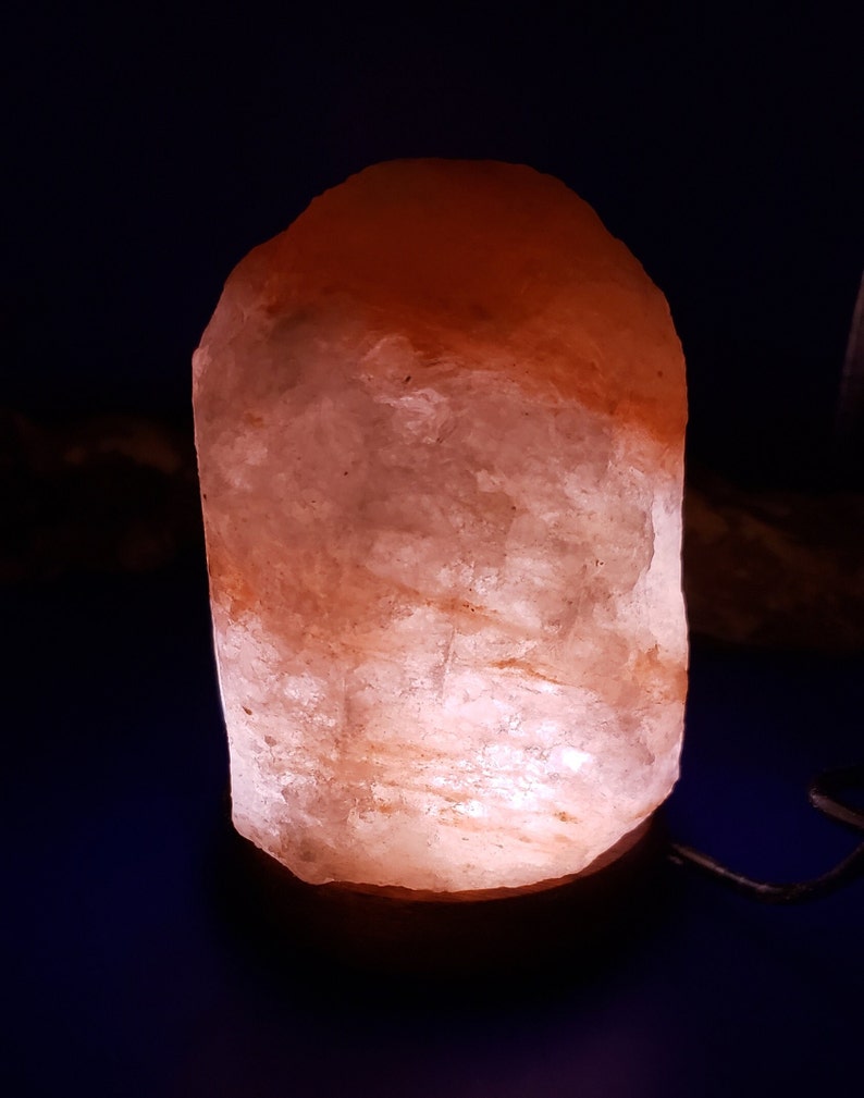 K&ouml;nnte beinhalten: Eine rosa Himalaya-Salzlampe mit einem Holzsockel. Die Lampe ist eingeschaltet und leuchtet.