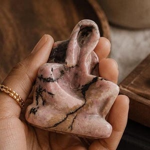 May include: A pink and black stone figurine of a seated person. The figurine is held in a hand, with a gold ring on a finger. A candle in a glass jar and a pair of eyeglasses are in the background.