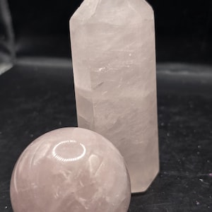 May include: A rose quartz crystal sphere and a rose quartz obelisk are displayed against a dark background. The sphere is smooth and round, while the obelisk is a tall, six-sided point. Both are a pale pink color.