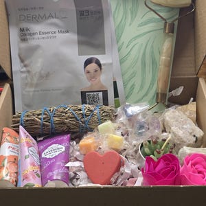 May include: A gift box filled with self-care items. Includes a face mask, jade roller, hand creams, sage bundle, heart-shaped soap, decorative flowers, and crystals. The box is lined with white paper.