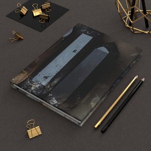 May include: A hardback journal with a dark, textured cover featuring a black crystal design. Gold binder clips and pencils are scattered around the journal on a dark gray surface. A gold geometric pen holder is in the background.