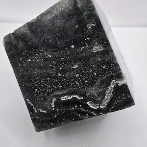 May include: A dark green, cube-shaped stone with a textured surface and white veining. The stone has a polished appearance and is angled slightly, showcasing its geometric form and natural patterns. The overall aesthetic is modern and minimalist.