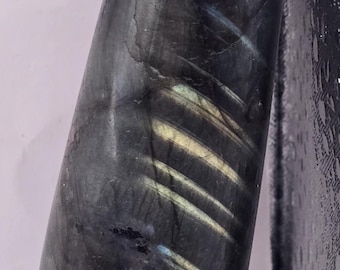 May include: A polished, elongated stone with a dark gray base color and iridescent flashes of blue, yellow, and green. The stone has a smooth, rounded shape and appears to be a decorative or healing crystal.