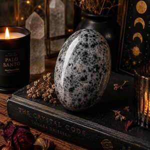 May include: A speckled gray and white egg-shaped stone sits atop a dark book titled "The Crystal Code" by Tamara Driessen. A lit black candle and dried flowers are in the background, creating a mystical aesthetic.