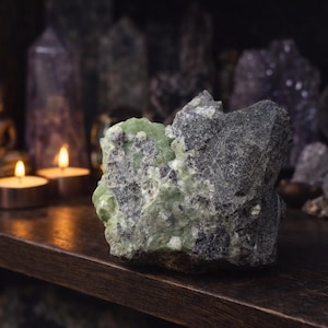 May include: A rough-hewn, natural stone specimen with a striking contrast of green and gray hues. The mineral sits on a dark wooden shelf, with a blurred background of other crystals and a small Buddha statue. Two lit candles add a warm glow.