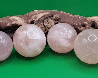 May include: Four smooth, polished rose quartz spheres, each with a slightly different shade of pink, are arranged in a row on a piece of driftwood. The spheres are all about the same size and have a natural, organic look.