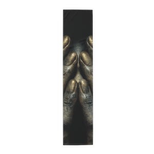 May include: A rectangular table runner with a close-up image of hands clasped in prayer. The hands are a muted gold colour, with detailed texture and shading. The background is solid black, creating a dramatic contrast.