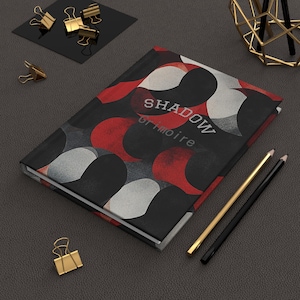 Op de afbeelding: Een hardcover journal met de woorden "SHADOW grimoire" op de cover. Het coverontwerp heeft een patroon van rode, witte en zwarte vormen. Gouden en zwarte potloden en gouden paperclips zijn ook zichtbaar.