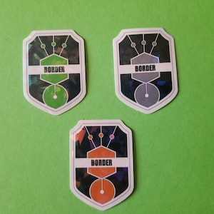 May include: Three shield-shaped stickers with a black and white geometric design. Each sticker has a different colored circle in the center: green, gray, and orange. The word "BORDER" is printed in white on each sticker.