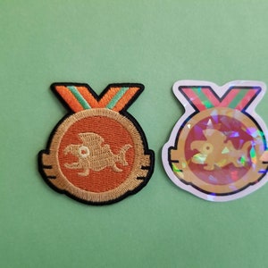 May include: Two embroidered patches of a gold fish with orange and green accents on a medal. One patch is on a green background and the other is on a white background.
