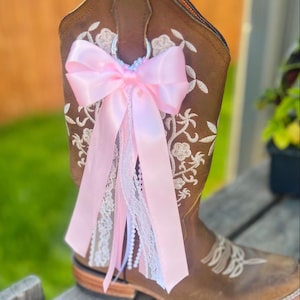 May include: A brown leather cowboy boot featuring white floral embroidery on the shaft. A decorative accessory is attached to the top pull tab, consisting of a large pink satin bow with cascading ribbons, white lace trim, and a string of white beads.