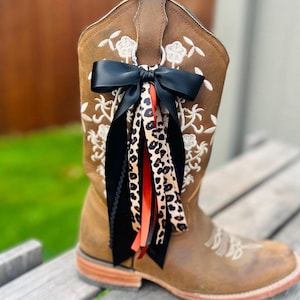 May include: A brown leather cowboy boot featuring white floral embroidery and a decorative accessory attached to the pull tab. The accessory consists of a black satin bow with hanging ribbons in black, solid orange, and a tan leopard print pattern.