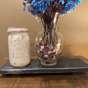 May include: A black wooden tray with a whitewashed finish sits on a wooden surface. A white mason jar with a twine lid and the text "Ball Wide Mouth" is on the tray. A clear glass vase with blue flowers and red and white glass stones is on the tray.