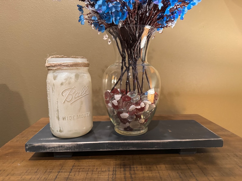 May include: A black wooden tray with a whitewashed finish sits on a wooden surface. A white mason jar with a twine lid and the text "Ball Wide Mouth" is on the tray. A clear glass vase with blue flowers and red and white glass stones is on the tray.