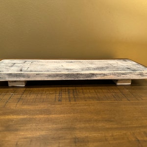 May include: A whitewashed wooden tray with a rectangular shape and slightly raised edges. The tray has two small wooden feet on each side, giving it a rustic look.