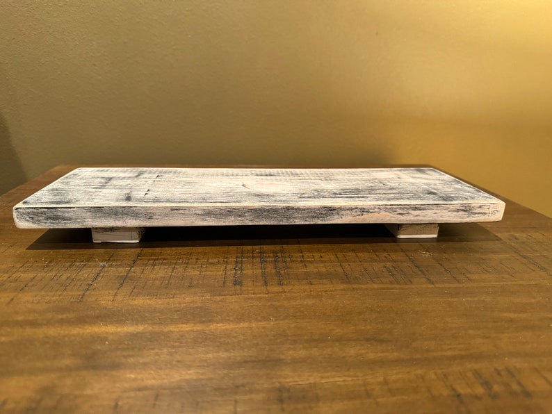 May include: A whitewashed wooden tray with a rectangular shape and slightly raised edges. The tray has two small wooden feet on each side, giving it a rustic look.