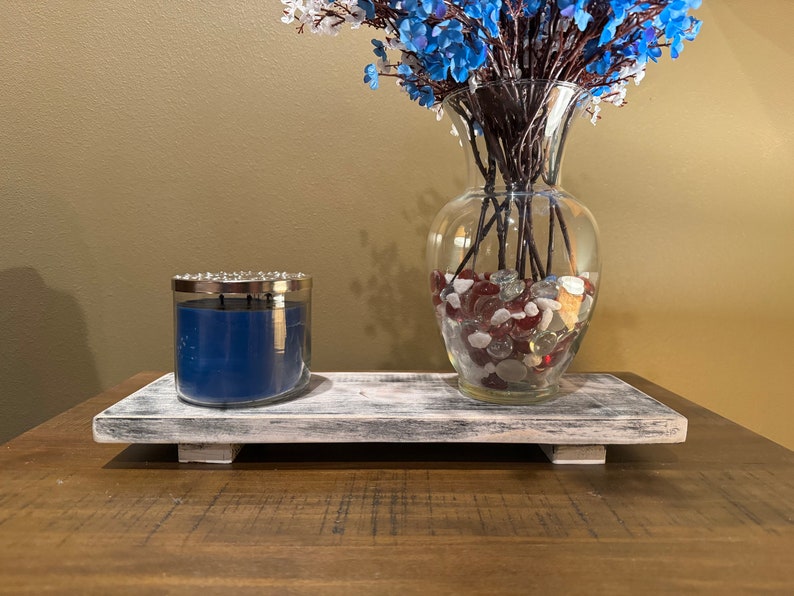 May include: A whitewashed wooden tray with a blue candle and a glass vase filled with blue flowers and red and white glass stones.