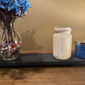 May include: A black wooden tray with a clear glass vase filled with red and white stones and blue flowers, a clear glass jar with a brown twine tied around the lid, and a blue candle with a silver lid.
