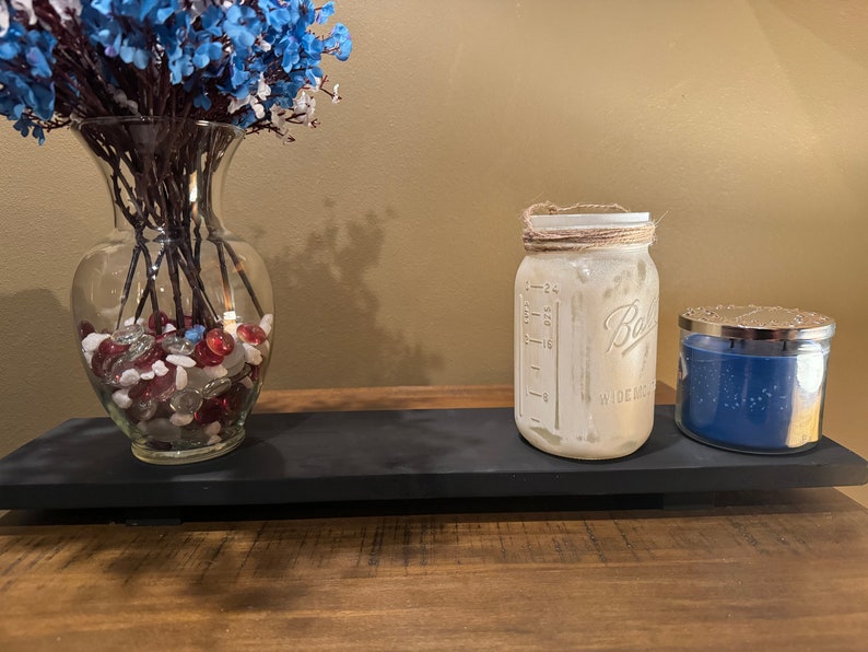 May include: A black wooden tray with a clear glass vase filled with red and white stones and blue flowers, a clear glass jar with a brown twine tied around the lid, and a blue candle with a silver lid.
