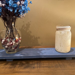 May include: A blue wooden shelf with a clear glass vase filled with red and white stones and a white mason jar with a brown twine lid. The vase holds blue and white flowers.