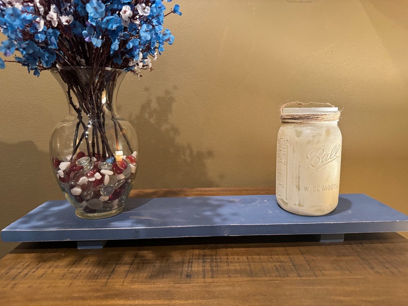 May include: A blue wooden shelf with a clear glass vase filled with red and white stones and a white mason jar with a brown twine lid. The vase holds blue and white flowers.
