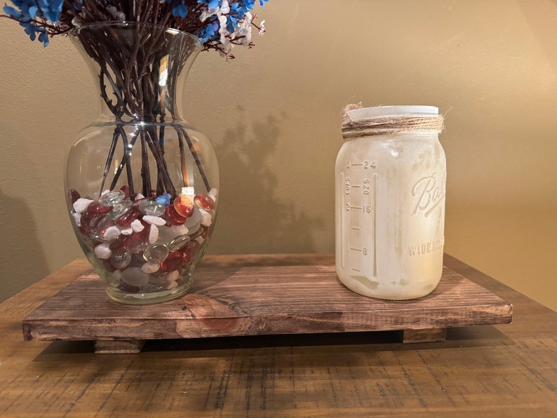 May include: A rustic wooden tray with a clear glass vase filled with red, white, and blue glass stones and a white mason jar with a twine lid. The mason jar has measurements in ounces and cups.