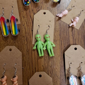 May include: Assortment of earrings featuring miniature skateboard, alien, and baby figures. The earrings are displayed on brown card stock.