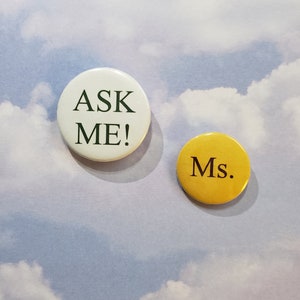 Może przedstawiać: Dwa okrągłe guziki z tekstem. Większy guzik jest biały z zielonym tekstem, który mówi "ASK ME!" Mniejszy guzik jest żółty z czarnym tekstem, który mówi "Ms."