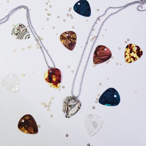 Op de afbeelding: Twee zilveren kettingen met gitaarplectrum hangers. De plectrums zijn gemaakt van verschillende materialen, waaronder schelp, hout en plastic. De kettingen liggen op een witte achtergrond met gouden glitter.