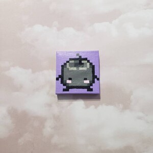 Puede incluir: Un pequeño lienzo pintado a mano con un personaje pixelado con cuerpo gris, ojos negros y mejillas rosadas. El personaje está sobre un fondo morado claro.