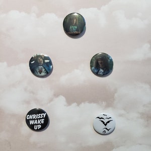 May include: Five round button pins with different designs. One pin is black with white text that reads "Chrissy Wake Up". One pin is white with a black silhouette of bats. The remaining three pins feature black and white photos of a person with long hair.
