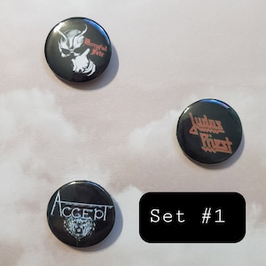 Może przedstawiać: Zestaw trzech czarno-białych pinów z logo zespołów. Piny przedstawiają logo Accept, Judas Priest i Mercyful Fate. Tekst "Set #1" jest nadrukowany na czarnym prostokącie.