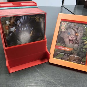 Könnte beinhalten: Eine rote Kartenaufbewahrungsbox aus Kunststoff gefüllt mit einem Kartendeck und ein separater orangefarbener Kunststoff-Displayrahmen mit einer einzelnen Karte, die eine Kreatur und den Text Pridestalker sowie Spielstatistiken zeigt.
