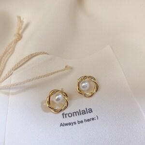 May include: A pair of gold-toned earrings with a pearl in the centre of each earring. The earrings are in the shape of a circle with a twisted design. The text "fromlala Always be here:" is printed on the white surface below the earrings.