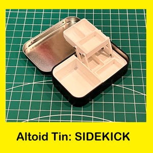May include: A white plastic organizer with multiple compartments, folded open inside a silver Altoids tin. The tin is labeled "Altoid Tin: SIDEKICK".