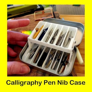 May include: A metal calligraphy pen nib case with a white interior, holding various nibs in different shapes and colors. The case is open, revealing the nibs, and the text "Calligraphy Pen Nib Case" is at the bottom.