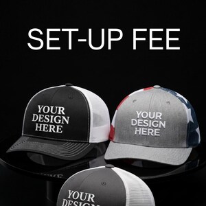 May include: Three trucker hats with the text "YOUR DESIGN HERE" in white. One hat is black with a white mesh back, another is gray with a white mesh back, and the third is black with a white mesh back. The text "SET-UP FEE" is at the top.