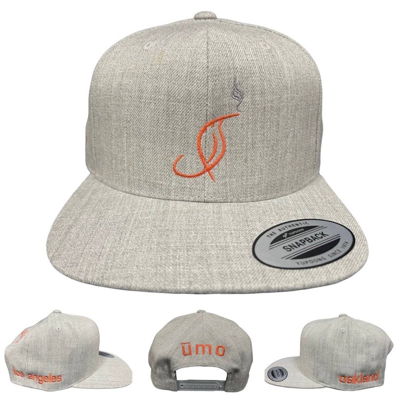 Puede incluir: Gorra snapback gris claro con un dise&ntilde;o bordado en naranja de una letra "J" estilizada con humo. La gorra tiene una visera plana y las palabras "los angeles" y "oakland" en naranja. La parte trasera de la gorra tiene la palabra "umo" en naranja.