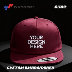 Może przedstawiać: Bordowa czapka z daszkiem typu snapback z białym haftowanym napisem "YOUR DESIGN HERE". Czapka ma płaski daszek i etykietę "SNAPBACK". Słowa "CUSTOM EMBROIDERED" znajdują się na dole.