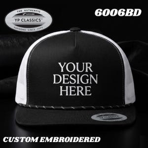 Puede incluir: Una gorra de camionero negra y blanca con visera plana, un detalle de cuerda en la base de la corona y paneles laterales de malla blanca. El panel frontal muestra el texto bordado YOUR DESIGN HERE. Una pegatina YP Classics 6006BD está en la visera.