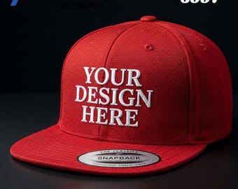 May include: A red baseball cap with a flat brim and a snapback closure. The front features white embroidered text that reads YOUR DESIGN HERE. A circular sticker on the brim displays the text THE CLASSICS SNAPBACK YUPOONG SINCE 1974.