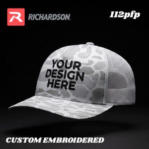 May include: A gray and white camouflage pattern baseball cap with a mesh back. The cap features the text "YOUR DESIGN HERE" in black, and the words "CUSTOM EMBROIDERED" are at the bottom.