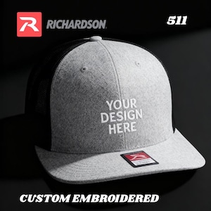 May include: A gray Richardson 511 trucker hat with a black mesh back and a curved brim. The front of the hat features the text "YOUR DESIGN HERE" in white. The words "CUSTOM EMBROIDERED" are at the bottom.
